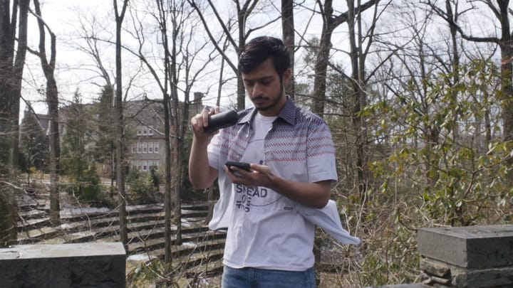 Harsha is standing in front of a forest, holding a speaker and looking at his phone.