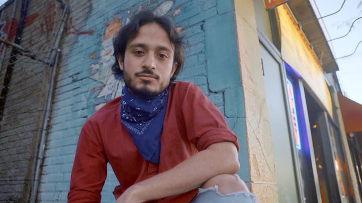 Harsha, a man, is squatting in front of a painted wall wearing a red shirt and blue bandanna.